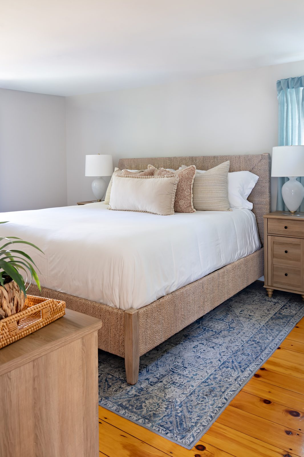 Indian Road coastal bedroom with woven bed frame, white bedding, blue area rug, and pine floors